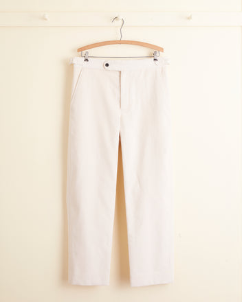 A pair of Winter Farm Trousers in light beige corduroy is neatly hanging on a wooden hanger against a plain cream-colored wall.