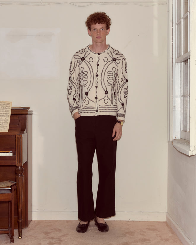 A person with curly hair stands indoors wearing the Winter Garden Cardigan, paired with black pants and black shoes.