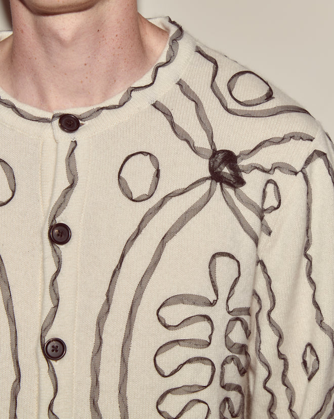 Close-up of the Winter Garden Cardigan, a cream knitwear piece featuring black abstract line designs and dark buttons, worn by a person.