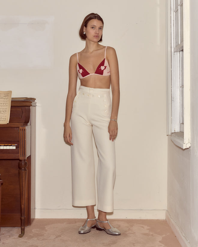 A woman in white high-waist pants and the Wonderland Bra with a red hearts motif stands indoors by a piano and window, evoking whimsical Alice Through the Looking Glass vibes.