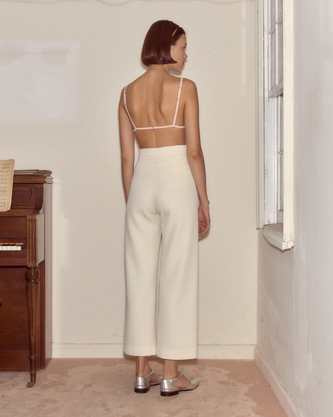 A woman in the Wonderland Bra and white pants stands facing a corner of a minimal room, sunlight streaming beside a piano—evoking quiet curiosity reminiscent of Alice Through the Looking Glass.