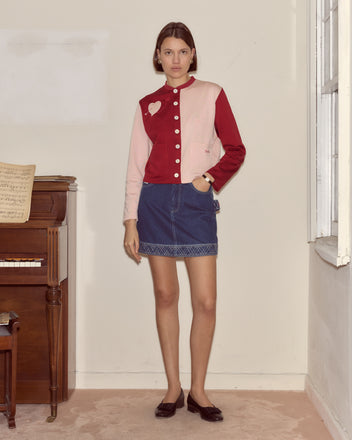 Wearing the Wonderland Cardigan with a denim skirt, a woman stands by a piano in soft light, her look capturing the whimsical charm of Alice through the Looking Glass.
