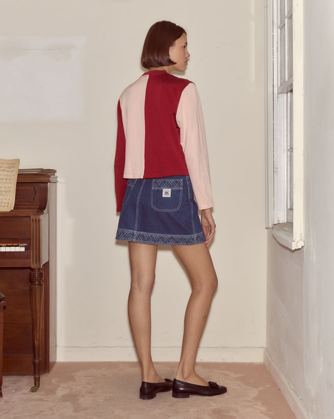A woman stands indoors facing away, wearing the Wonderland Cardigan with a red and pink hearts motif and a denim skirt near a piano and window.