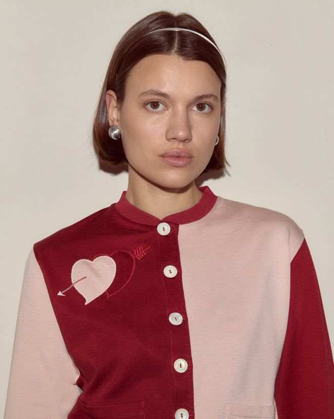 Woman with short brown hair wears the Wonderland Cardigan in red and pink, featuring a heart and arrow motif on the chest, reminiscent of whimsical knitwear from Alice Through the Looking Glass.