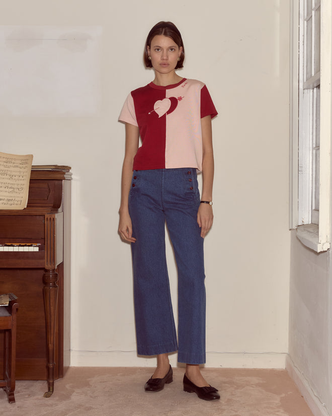 A woman stands indoors by a piano, wearing the Wonderland Tee inspired by Alice Through the Looking Glass, paired with blue jeans and black shoes.
