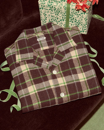 A folded Woodcrest Overshirt in brown and green flannel with cream checks rests next to a wrapped gift tied with a green ribbon.
