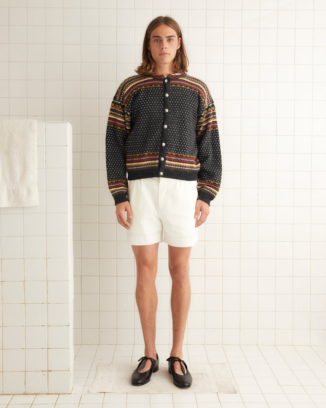 A person stands against a tiled wall wearing the Woodford Jacquard Cardigan, white shorts, and black shoes.