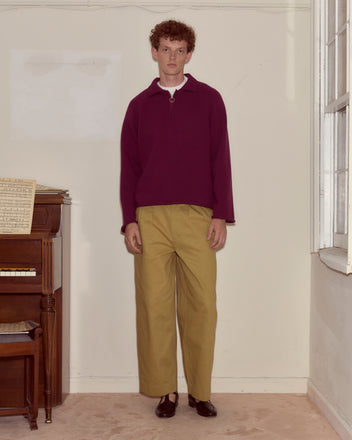 Someone wearing the Wool Dock Pullover, a chunky maroon oversized sweater, and tan pants stands indoors beside a piano and a large window.