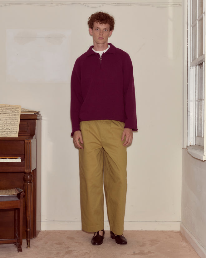 Someone wearing the Wool Dock Pullover, a chunky maroon oversized sweater, and tan pants stands indoors beside a piano and a large window.