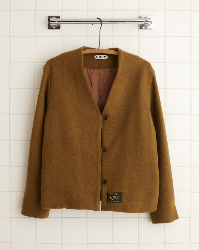 The Wool Drab Jacket - 4, featuring a distinctive textile, is elegantly draped on a hanger against a white tiled wall.