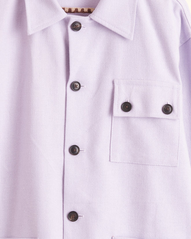 A Lilac Worker Jacket - S/M, featuring a light purple hue with black buttons and multiple pockets that embody a classic workwear silhouette, hangs on a hanger against a white background.