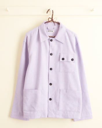 A Lilac Worker Jacket - S/M, featuring a light purple hue with black buttons and multiple pockets that embody a classic workwear silhouette, hangs on a hanger against a white background.