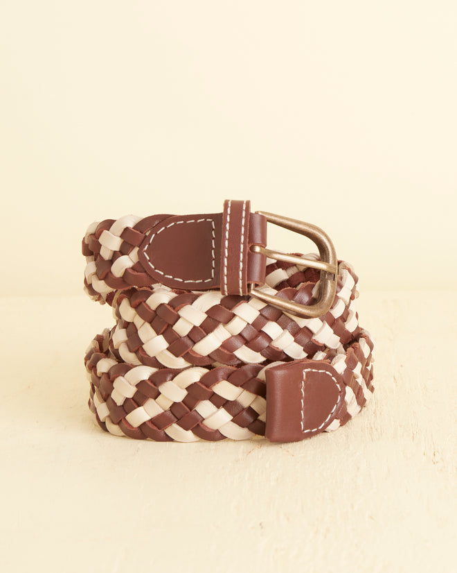 The Woven Belt - Brown/White, crafted with vintage style in brown and white leather and accented by a silver buckle, is elegantly coiled on a light surface.