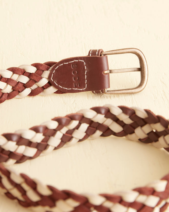 The Woven Belt - Brown/White, crafted with vintage style in brown and white leather and accented by a silver buckle, is elegantly coiled on a light surface.