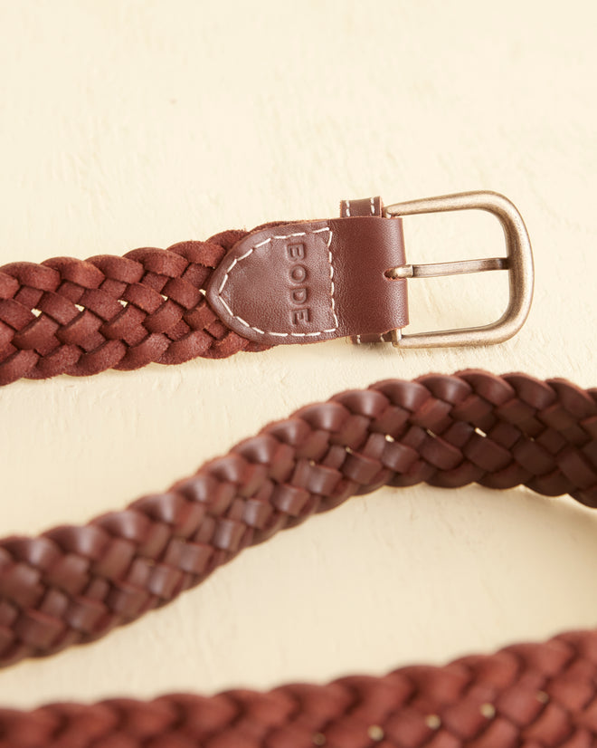 The Woven Belt - Brown, featuring a braided leather design with a metal buckle, reminiscent of vintage styles, stands coiled and upright on a light surface.