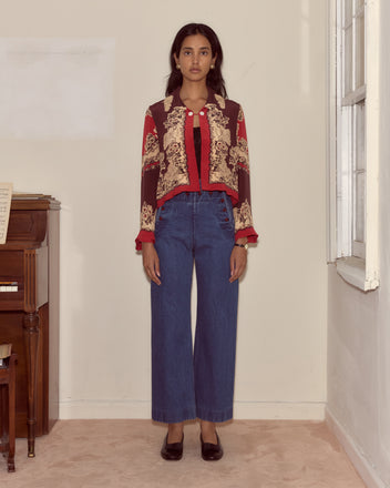A woman wearing a patterned red jacket, Denim Sailor Trousers in midweight denim, and black shoes stands by a piano and window.