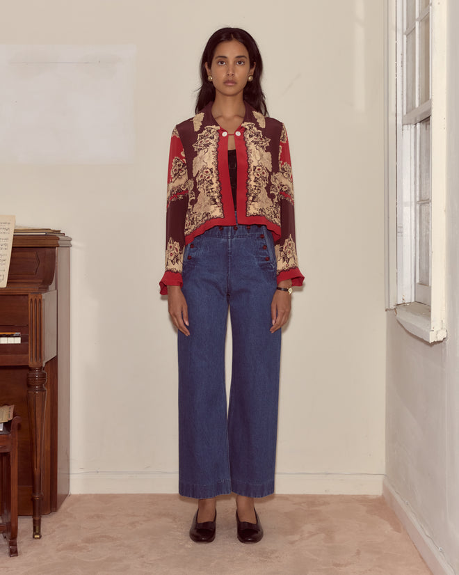 A woman wearing a patterned red jacket, Denim Sailor Trousers in midweight denim, and black shoes stands by a piano and window.