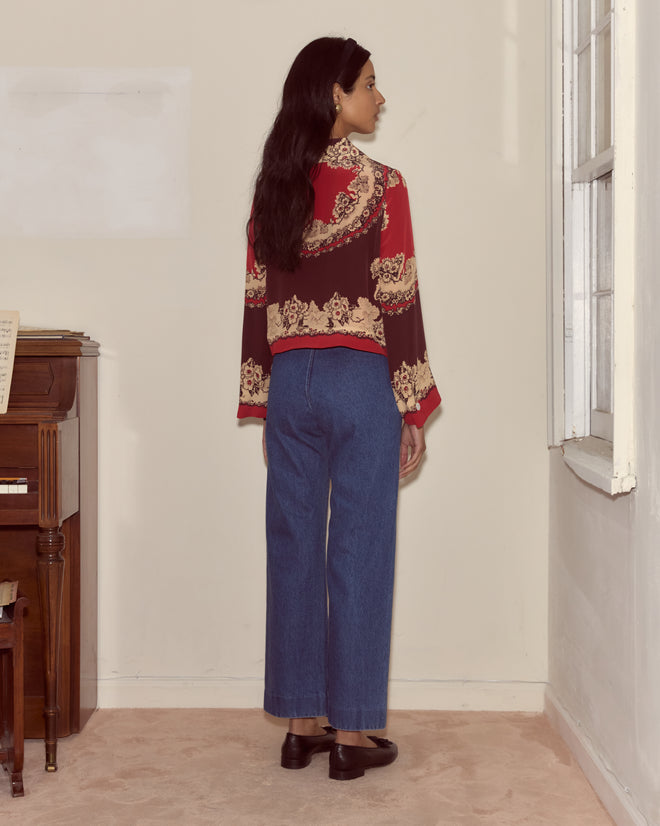 A woman with long dark hair stands facing a corner, wearing a patterned red top and classic Denim Sailor Trousers crafted from midweight denim.