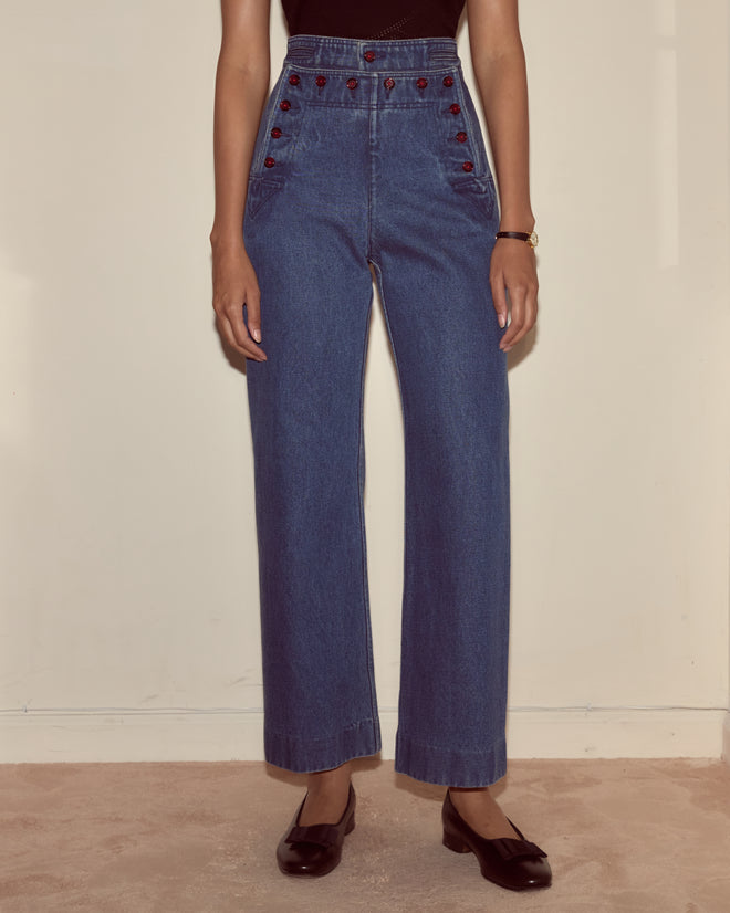 A person wearing Denim Sailor Trousers with red buttons, a black top, and black shoes, standing indoors.