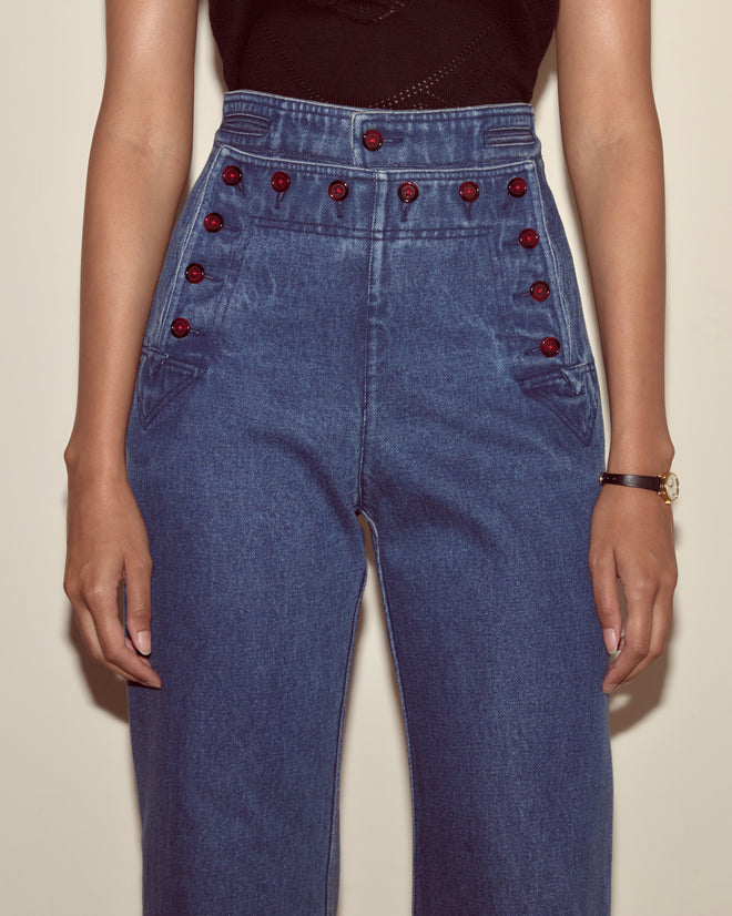 Person wearing Denim Sailor Trousers—high-waisted blue jeans with red buttons, made from 100% cotton—paired with a black sleeveless top, arms at sides.