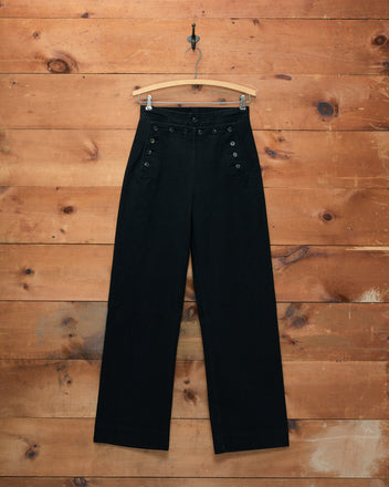 Denim Sailor Trousers - Black, high-waisted with wide legs and button details, displayed on a wooden hanger against a wood wall.