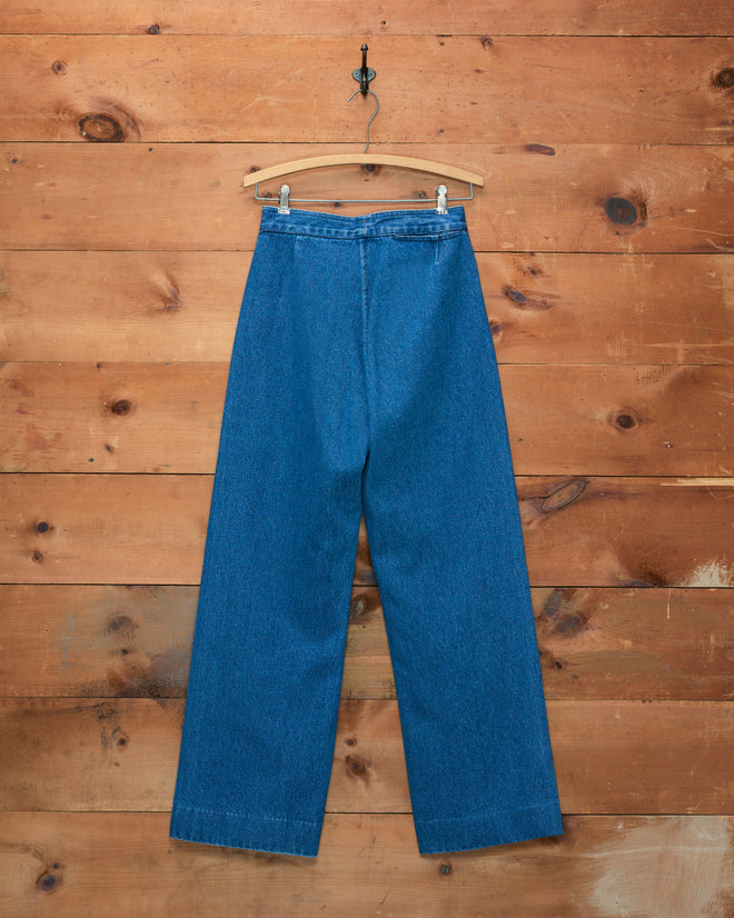Denim Sailor Trousers in a blue wash, featuring a high-waisted wide-leg silhouette and crafted from midweight denim, hang elegantly on a hanger against a wooden plank wall background.