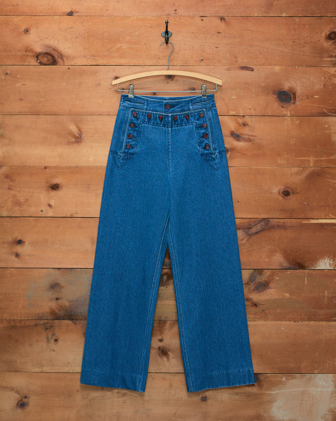 Denim Sailor Trousers crafted from 100% cotton midweight denim in blue, featuring a high-waisted fit, wide legs, and distinctive button details, are displayed hanging on a wooden wall.
