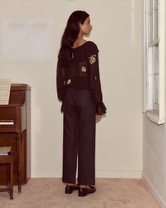 A woman wearing black Linen Sailor Trousers and a floral sheer blouse stands facing a wall near a piano and window.