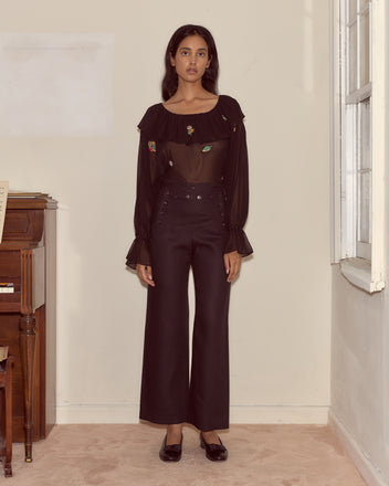 A woman stands indoors near a piano and window, wearing a sheer black blouse paired with Linen Sailor Trousers - Black.