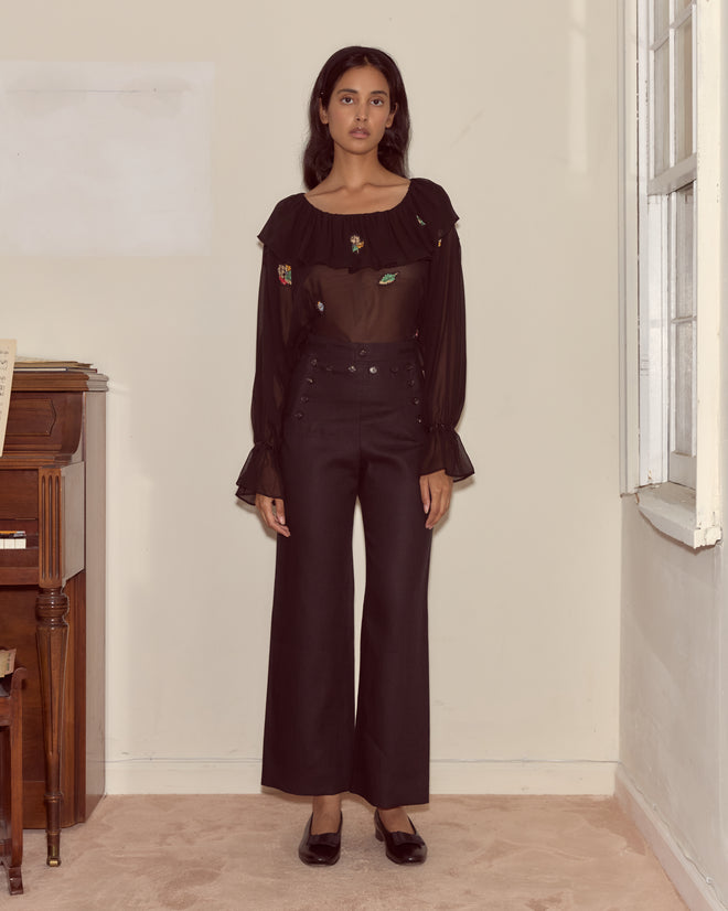 A woman stands indoors near a piano and window, wearing a sheer black blouse paired with Linen Sailor Trousers - Black.