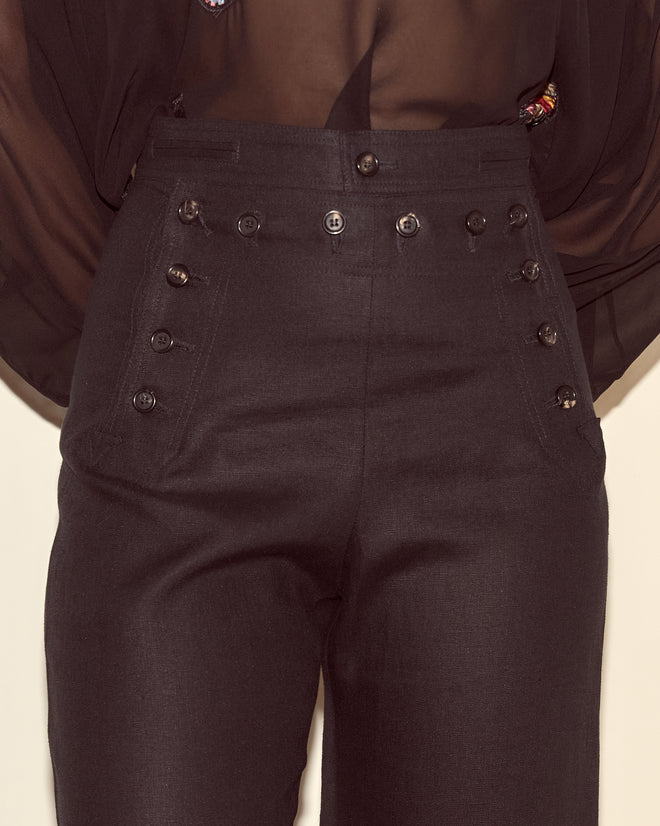 Close-up of high-waisted Linen Sailor Trousers in black, featuring multiple buttons, paired with a sheer black blouse.