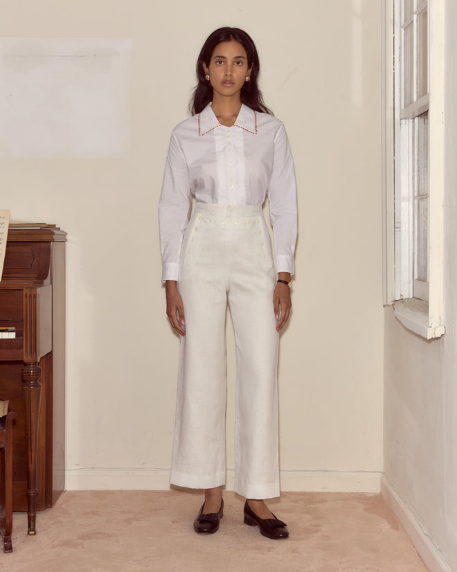 A woman in a white blouse and Linen Sailor Trousers - White stands indoors by a piano and window, looking at the camera.