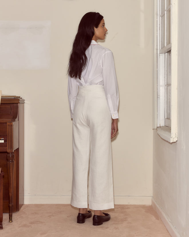 Wearing Linen Sailor Trousers - White, a woman stands by a cream wall near a window and piano, evoking 1940s style.