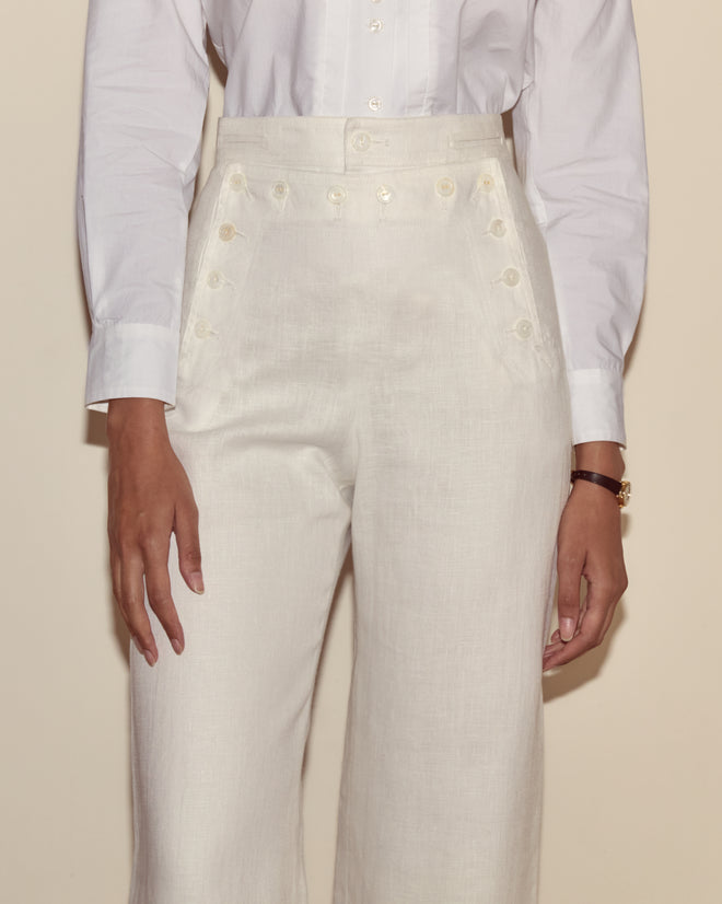 A woman wears Linen Sailor Trousers in white, featuring a high-waisted, 1940s-inspired design with button details, paired with a crisp white long-sleeve shirt.