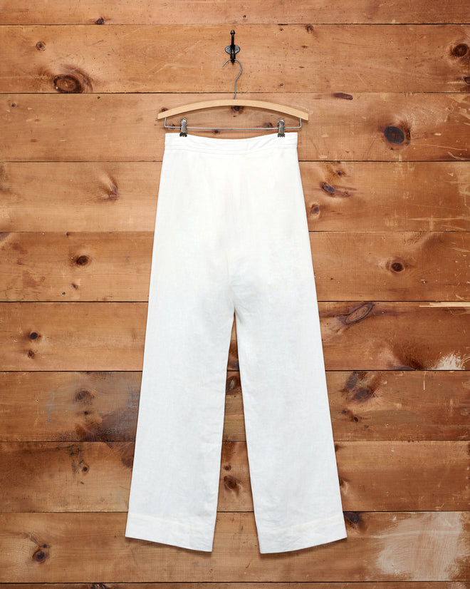 Linen Sailor Trousers - White hanging on a wooden hanger against a rustic wooden plank wall.