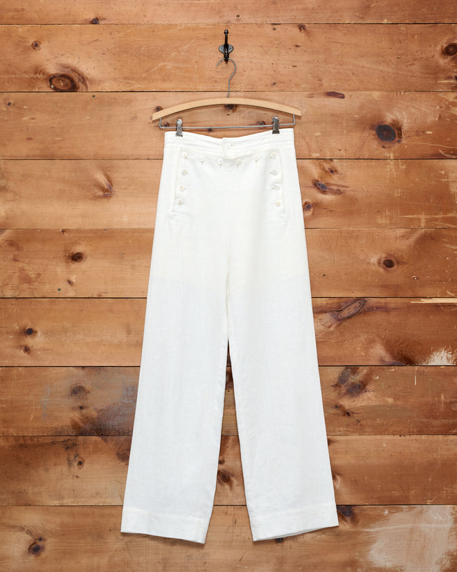 Linen Sailor Trousers - White, a pair of 1940s inspired high-waisted white linen trousers with button details, hanging on a wooden hanger against a wood wall.