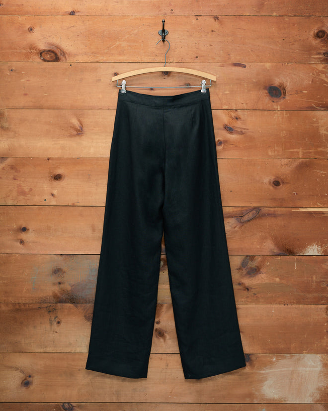 The Linen Sailor Trousers - Black, featuring a wide-leg silhouette and displayed on a hanger against a wooden wall background, evoke a classic 1940s style.