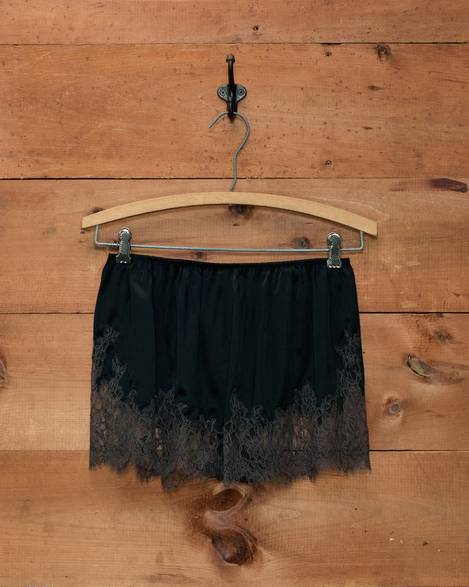 Black Lace Inlay Tap Shorts with a scalloped lace hem hanging on a wooden hanger against a wood wall.