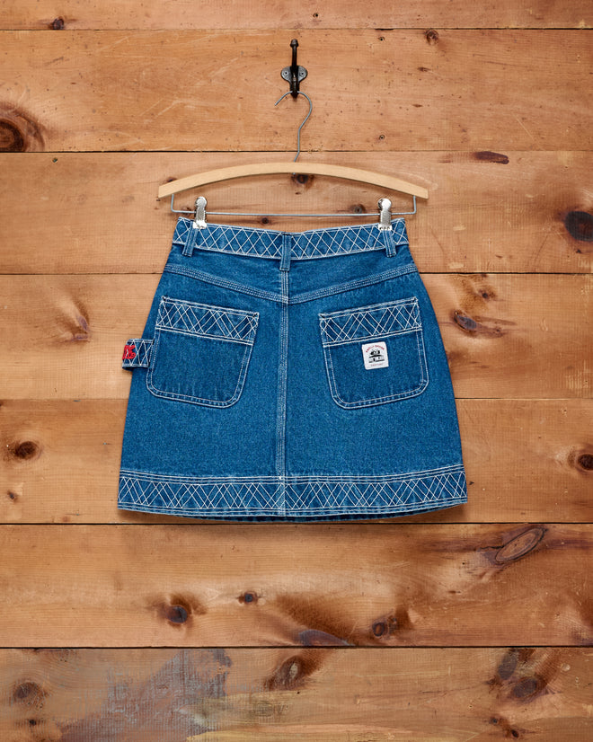 Embroidered Denim Skirt - Indigo, featuring white stitching details, is shown hanging on a wooden wall by a hanger; this classic piece adds a touch of workwear style to any outfit.