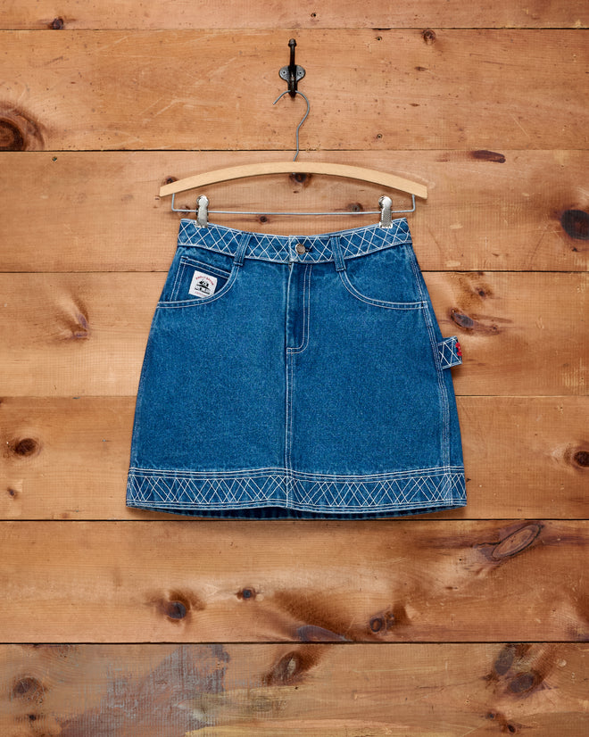The Embroidered Denim Skirt - Indigo, featuring white stitching, hangs on a wooden hanger against a wooden wall background, showcasing the timeless appeal of classic workwear style.