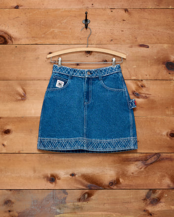 The Embroidered Denim Skirt - Indigo, featuring white stitching, hangs on a wooden hanger against a wooden wall background, showcasing the timeless appeal of classic workwear style.