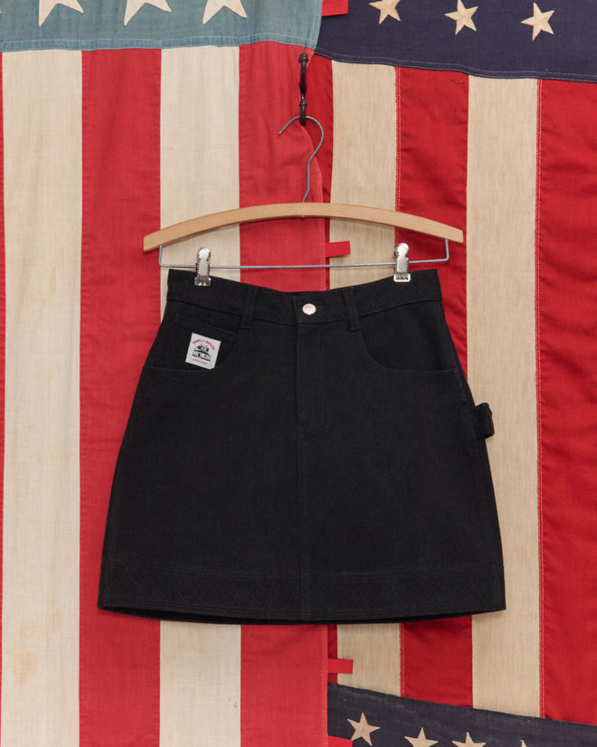 The Embroidered Denim Skirt - Black hangs on a wooden hanger with workwear details, set against a vintage American flag backdrop.