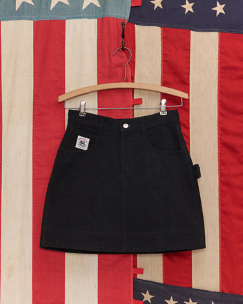 The Embroidered Denim Skirt - Black hangs on a wooden hanger with workwear details, set against a vintage American flag backdrop.