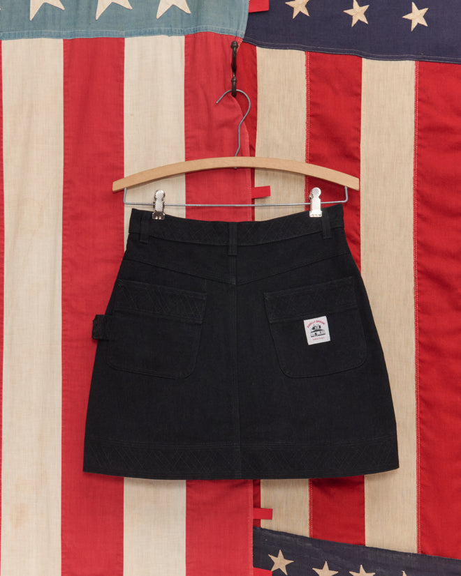 The Embroidered Denim Skirt - Black, featuring front pockets and workwear accents, hangs on a wooden hanger with an American flag in the background.