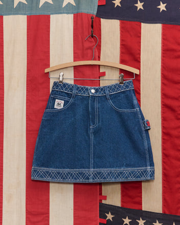 The Embroidered Denim Skirt with white stitching hangs on a wooden hanger against an American flag backdrop, highlighting its classic workwear style.