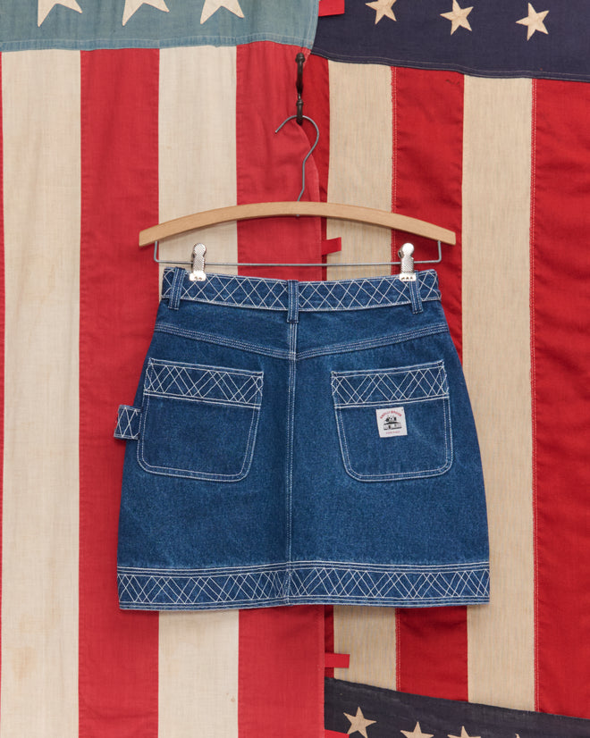 The Embroidered Denim Skirt, featuring white stitching, hangs on a hanger against an American flag backdrop, evoking classic workwear style.