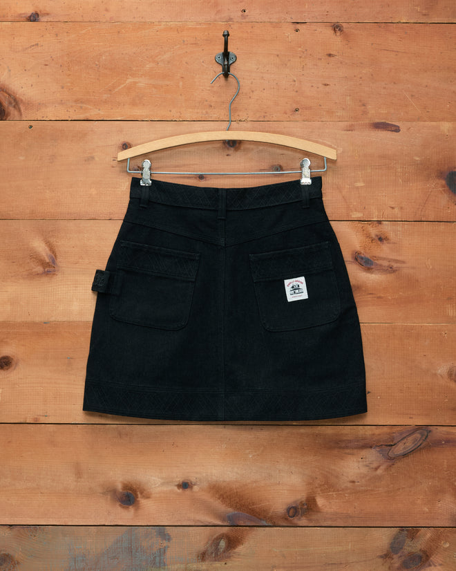 The Embroidered Denim Skirt - Black, featuring front pockets and subtle workwear details, hangs on a wooden hanger against a wooden wall background.