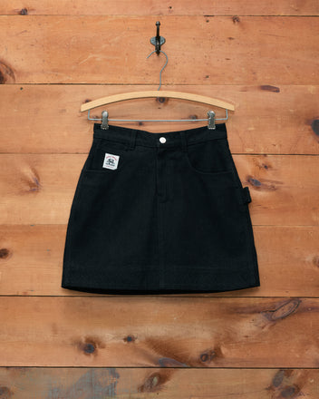 Embroidered Denim Skirt - Black with pockets and subtle workwear-inspired details, featuring zig-zag embroidery, displayed on a wooden hanger against a wooden wall background.