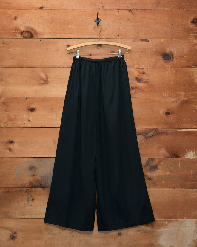 Roy Pants - Black wide-leg beach pants hanging on a wooden hanger against a rustic wooden wall background.