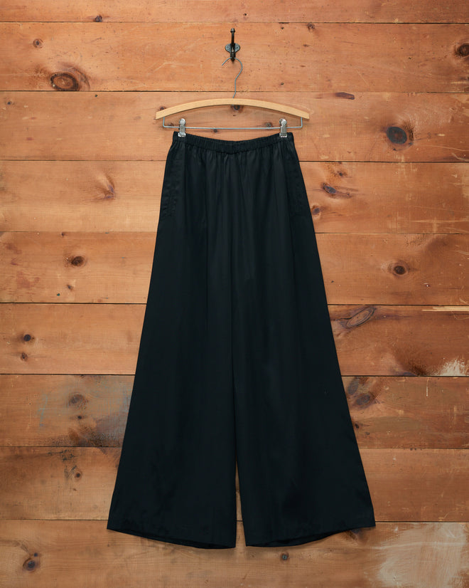Roy Pants - Black, featuring a wide-leg silhouette, displayed on a wooden hanger against a rustic wooden wall background.